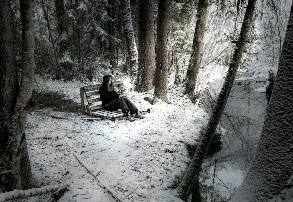 woman wearing black denim jeans sitting on bench surrounded by trees and covered with snow near body of water